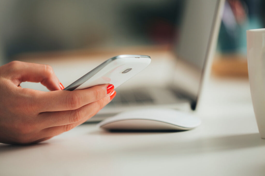 Woman's hand holding a smartphone with a laptop just behind the hand